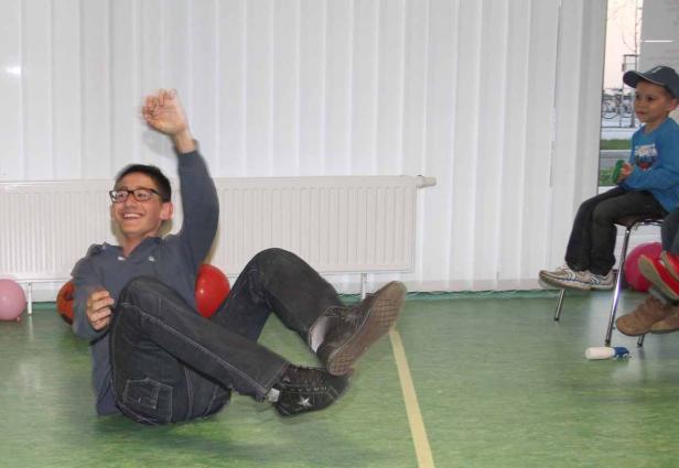 Ein Mann sitzt lachend auf dem Boden, während ein Junge im Hintergrund auf einem Stuhl sitzt.