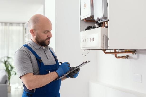 Handwerker in grauem Poloshirt und blauer Latzhose prüft Therme an der Wand und notiert auf Klemmbrett.