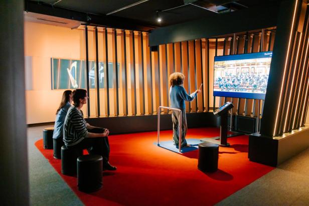 Eine Person steht in einem Raum mit einem Dirigierstab und spielt ein Spiel auf einem Bildschirm.