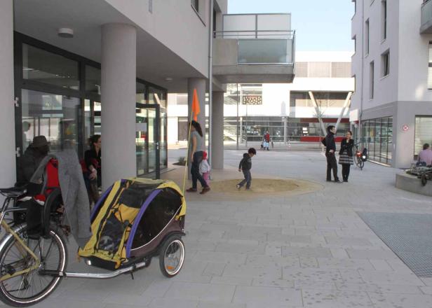 Ein Platz mit spielenden Kindern und Erwachsenen vor modernen Gebäuden.
