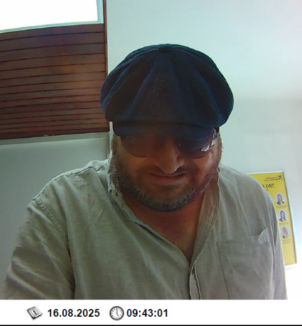 Ein Mann mit schwarzer Kappe und Sonnenbrille vor einem Bankomaten.