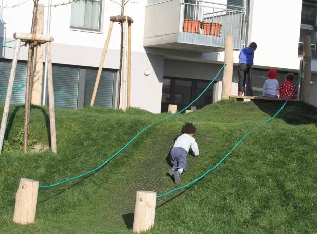 Kinder spielen auf einem grünen Hügel mit Seilen und Holzpfosten.