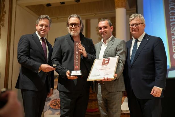 Vier Männer in Anzügen stehen zusammen, einer hält eine Trophäe und eine Urkunde, alle lächeln in festlicher Umgebung.