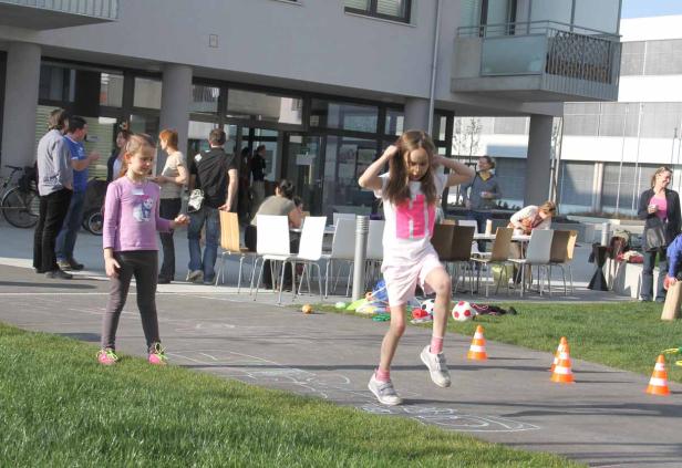 Ein Mädchen spielt im Freien Hüpfspiele, während andere Leute im Hintergrund stehen.