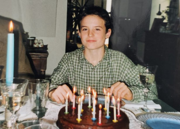 Jugendlicher sitzt vor Geburtstagskuchen mit brennenden Kerzen an festlich gedecktem Tisch.
