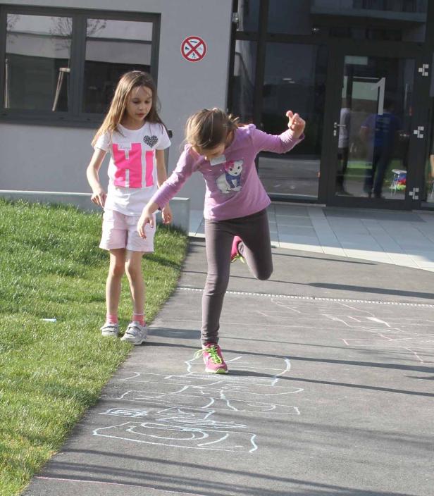 Zwei Mädchen spielen im Freien auf einem Gehweg mit Kreidezeichnung ein Hüpfspiel.