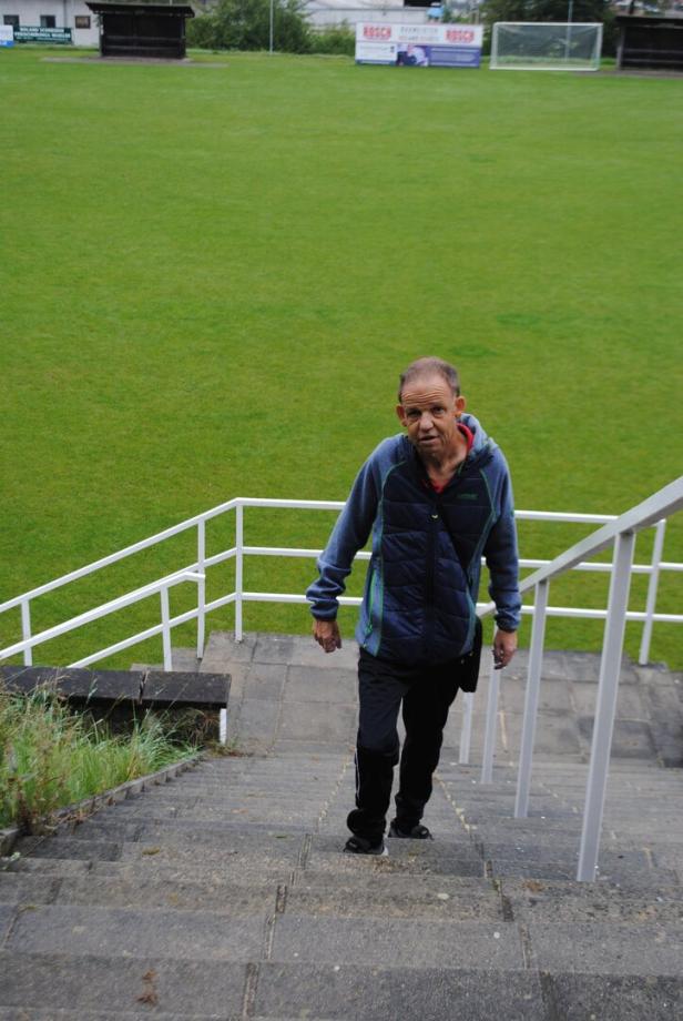Ein Mann mit Jacke geht eine Treppe neben einem Fußballfeld mit grünem Rasen hinauf.