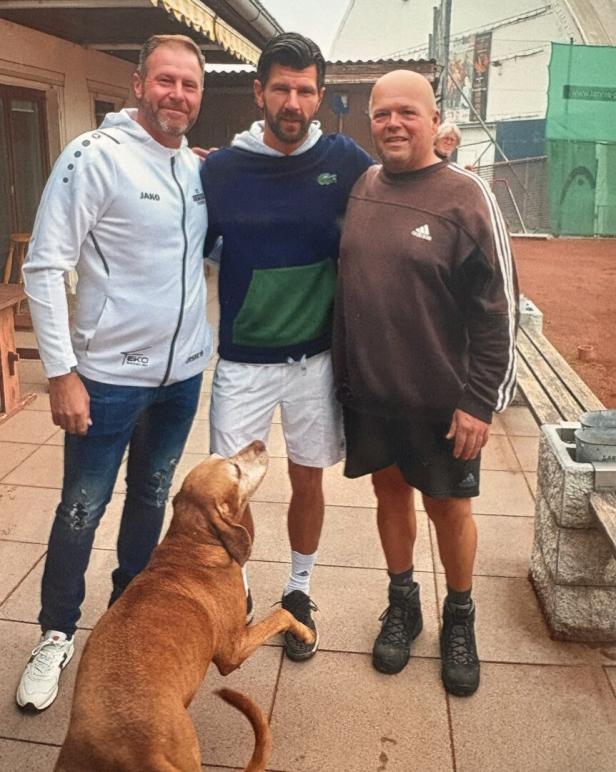 Drei Männer stehen nebeneinander auf einer Terrasse, ein brauner Hund hebt die Pfote zu einem von ihnen.