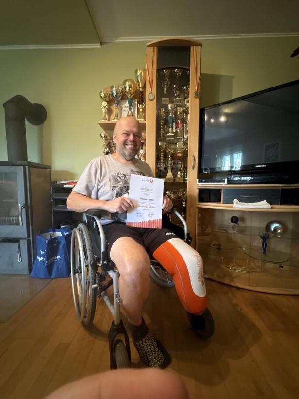 Ein Mann im Rollstuhl mit einer Beinprothese hält eine Urkunde und sitzt vor einer Vitrine voller Pokale und Medaillen.