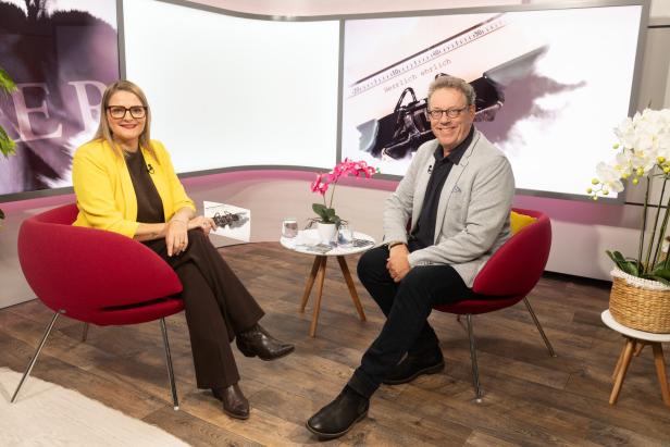 Zwei Personen sitzen einander in roten Sesseln gegenüber, zwischen ihnen ein kleiner Tisch mit Blumen und Wassergläsern.