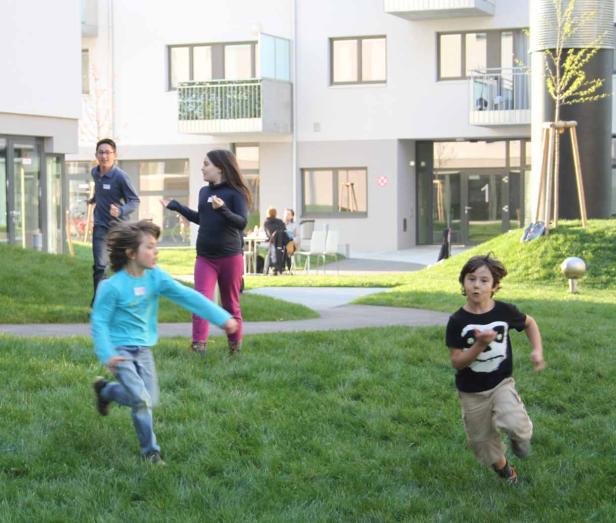 Zwei Kinder rennen auf einer Wiese vor einem modernen Wohngebäude.