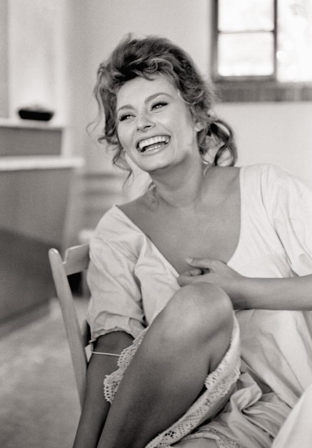 Sophia Loren sitzt entspannt auf einem Stuhl, hat eine Bluse an und lacht.
