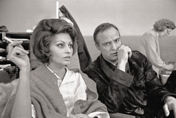 Am Set mit Marlon Brando. Beide Sitzen im Stuhl, sie raucht, er sieht ernst in die Kamera.