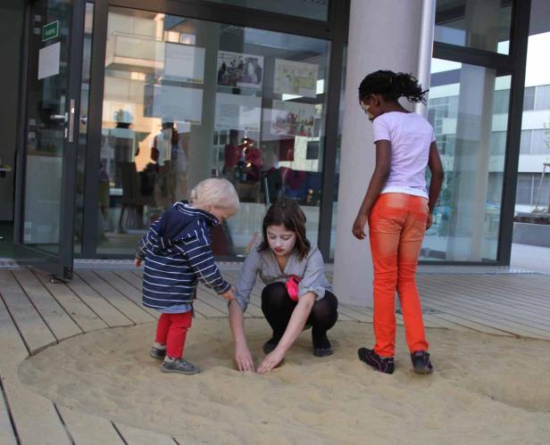 Drei Kinder spielen in einem Sandkasten vor einem Gebäude.