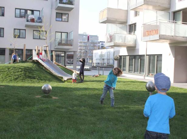 Kinder spielen auf einer Wiese mit Rutsche und Ball vor modernen Wohnhäusern.