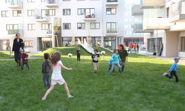 Kinder spielen auf einer Rasenfläche vor einem modernen Wohngebäude.