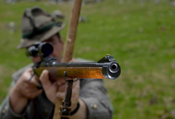 Eine Person mit Hut zielt mit einem Gewehr direkt nach vorne, der Lauf ist im Vordergrund scharf zu sehen.
