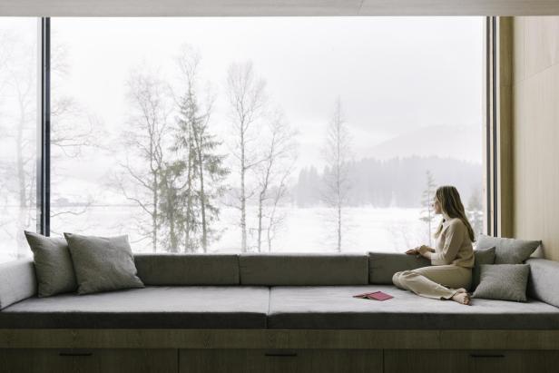 Eine Frau sitzt auf einer Couch in einem modernen Wohnzimmer und beobachtet die winterliche Landschaft durch eine große Glasfront.