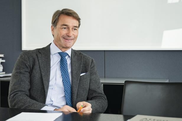 Ein Mann mit grauem Sakko und blauer Krawatte sitzt lächelnd an einem Tisch.