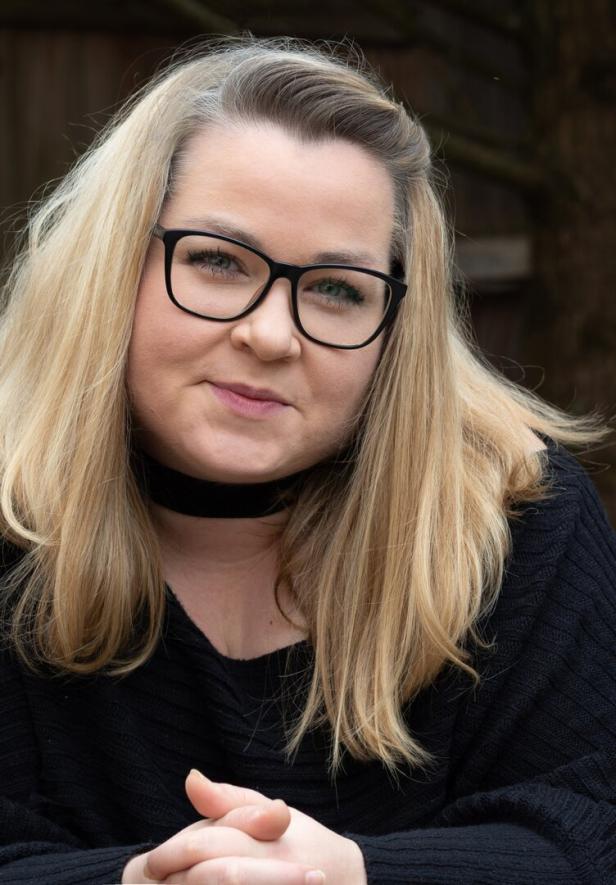 Autorin Julia Dippel mit langen blonden Haaren, schwarzer Brille und schwarzem Pullover sitzt draußen und lächelt leicht.