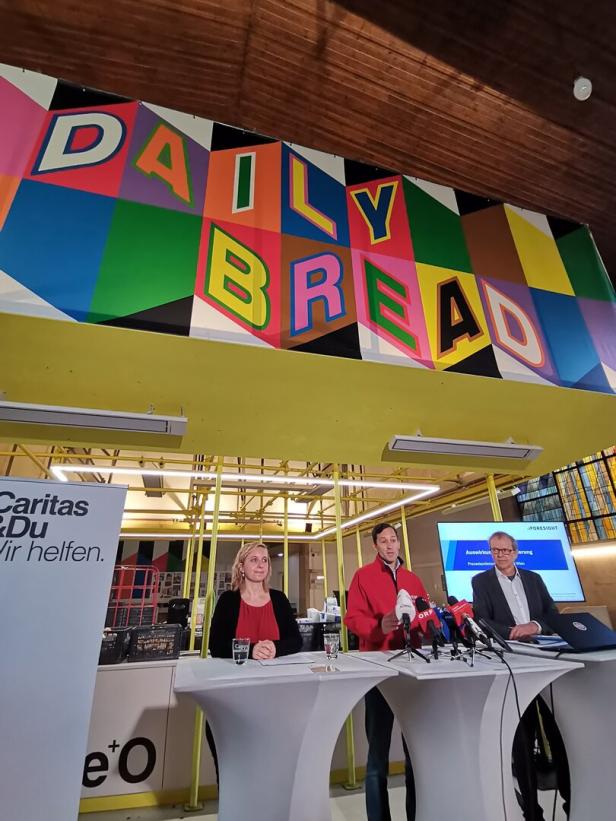 Lea Laubenthal, Caritasdirektor Klaus Schwertner und Christoph Hofinger bei einer Pressekonferenz, über ihnen eine Kunstinstallation, auf der Daily Bread steht..