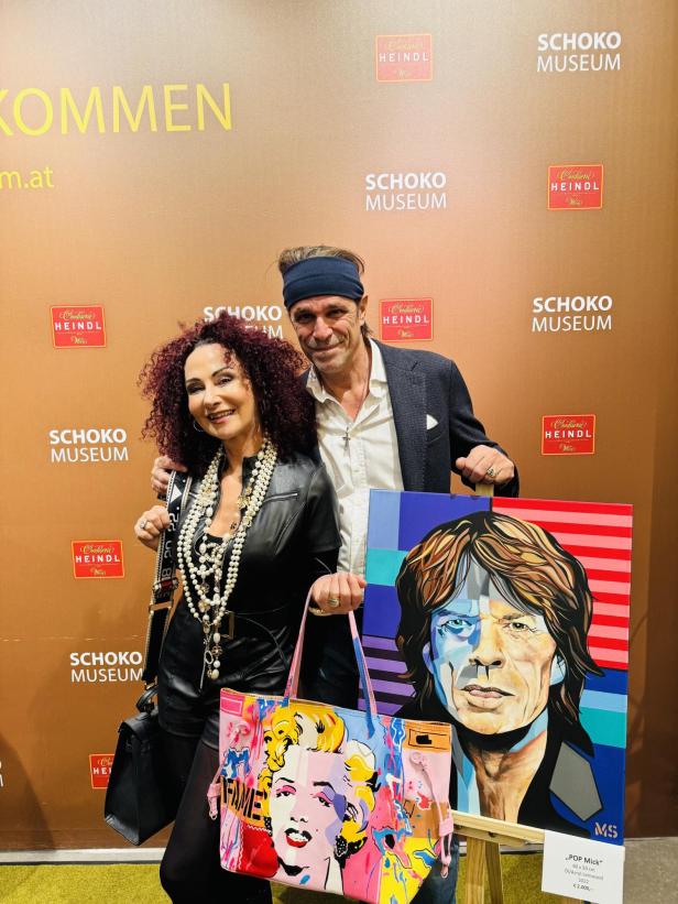 Zwei Personen posieren vor einer Wand, eine hält eine bunte Marilyn-Monroe-Tasche und ein Popart-Gemälde.