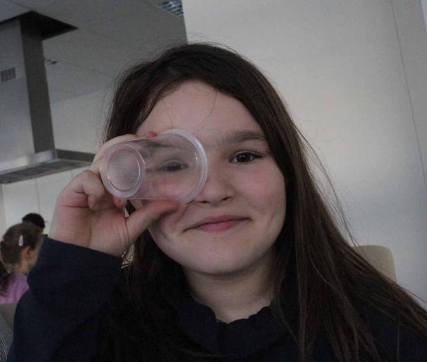 Ein Mädchen hält einen durchsichtigen Plastikbecher vor ihr Auge.