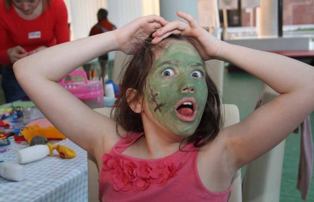 Ein Mädchen mit grün bemalten Gesicht und aufgerissenen Augen und Mund blickt in die Kamera.