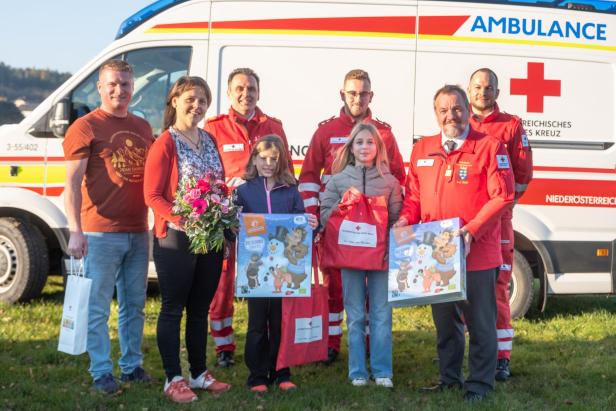 Eine Gruppe Menschen, darunter mehrere Männer in Rot Kreuz Uniform und zwei Mädchen mit Geschenken in ihren Armen, stehen vor einem Einsatzwagen und lächeln in die Kamera.