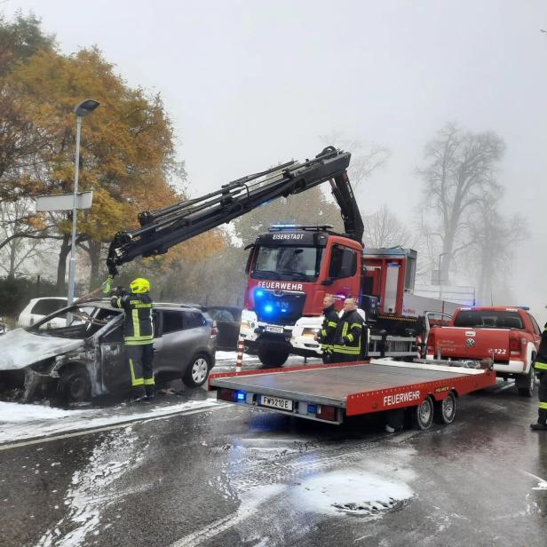 Feuerwehrleute bergen ein ausgebranntes Auto mit einem Kran, während Löschschaum auf der Straße liegt.