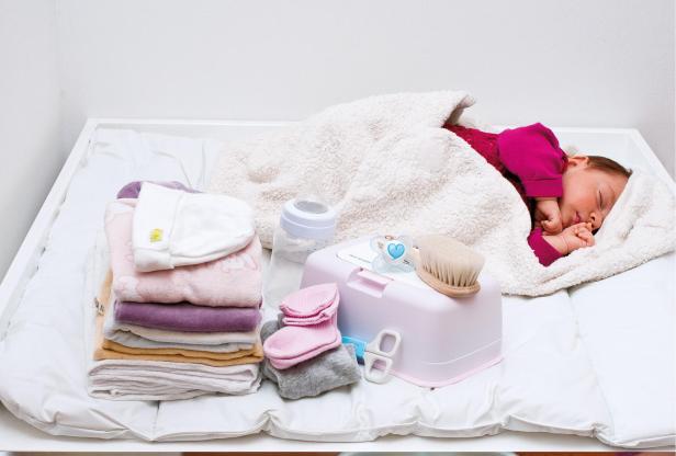 Schlafendes Baby in rotem Oberteil unter weißer Decke auf weißer Matratze, daneben Stapel bunter Babywäsche und Baby-Pflegeutensilien.