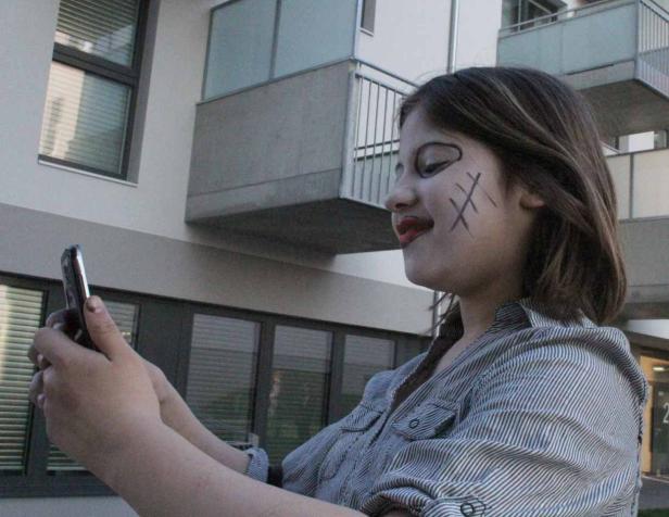 Ein Mädchen mit Gesichtsbemalung benutzt ein Smartphone vor einem Wohnhaus.