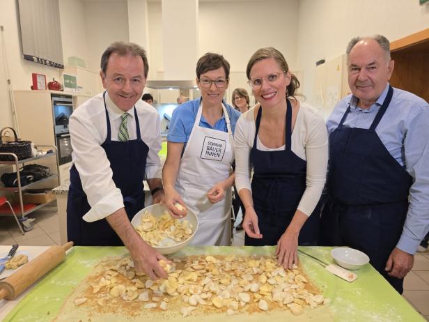 Vier Erwachsene bereiten gemeinsam in einer Küche einen Apfelstrudel zu und verteilen Apfelstücke auf Teig.