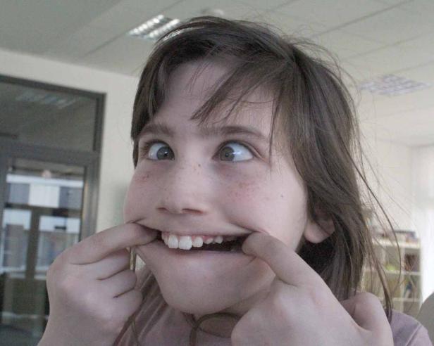 Ein Mädchen mit Sommersprossen verzieht das Gesicht und zieht mit den Fingern die Mundwinkel auseinander.