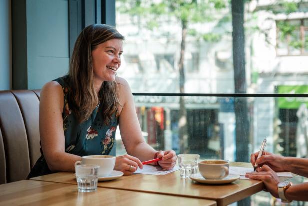 Eine Frau sitzt lächelnd an einem Tisch in einem Café, vor ihr stehen Tassen, Gläser und Papiere, sie unterhält sich mit einer anderen Person.