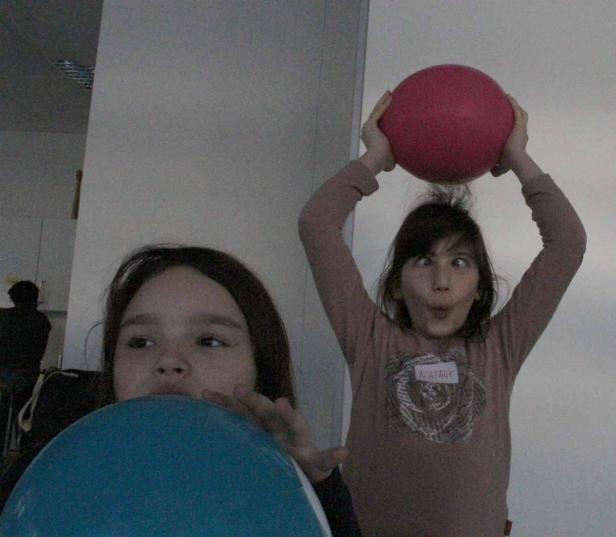 Zwei Mädchen spielen mit Luftballons, eines bläst einen blauen Ballon auf, das andere hält einen roten über den Kopf.