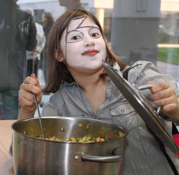 Ein geschminktes Mädchen rührt mit einem Löffel in einem großen Topf mit Reis.