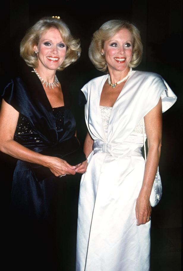 Zwei elegant gekleidete Frauen mit blonden Haaren stehen nebeneinander und lächeln, eine trägt Schwarz, die andere Weiß.