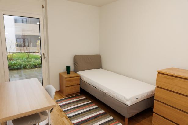 Ein schlicht eingerichtetes Zimmer mit Einzelbett, Nachttisch, Schreibtisch, Stuhl, Kommode und gestreiftem Teppich.