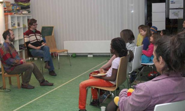 Eine Gruppe von Kindern und zwei Männer sitzen auf Stühlen in einem Raum.