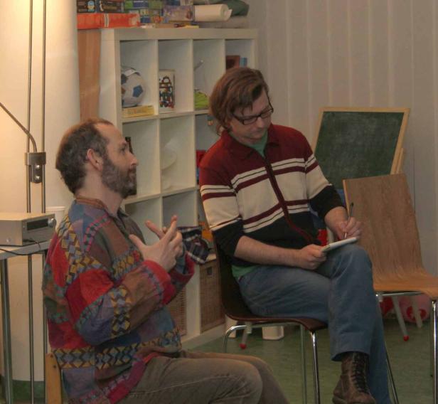 Zwei Männer sitzen in einem Raum, einer spricht, der andere macht Notizen.