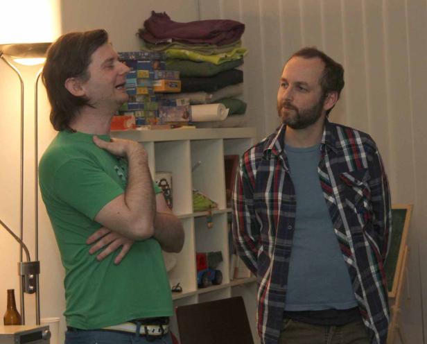 Zwei Männer unterhalten sich in einem Wohnzimmer vor einem Regal.