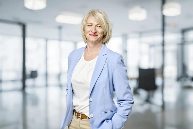 Frau mit blonden Haaren trägt hellblauen Blazer, weiße Bluse und beige Hose, steht mit einer Hand in der Hosentasche in einem modernen Büro.