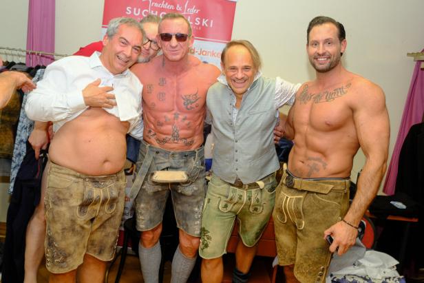 Vier Männer in Lederhosen, zwei davon oberkörperfrei mit Tattoos, stehen lachend nebeneinander in einem Raum.