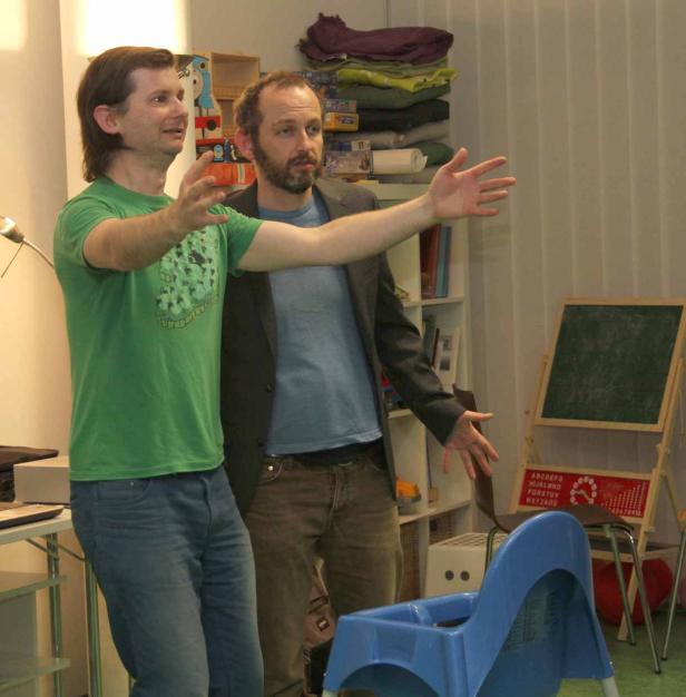 Zwei Männer gestikulieren in einem Raum mit einem Kinderstuhl und einer Tafel.