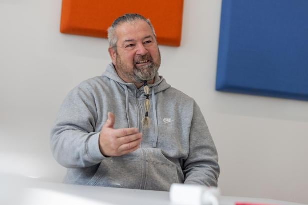 Ein Mann mit Bart und Zopf im grauen Nike-Hoodie gestikuliert vor orangefarbenen und blauen Wandelementen.