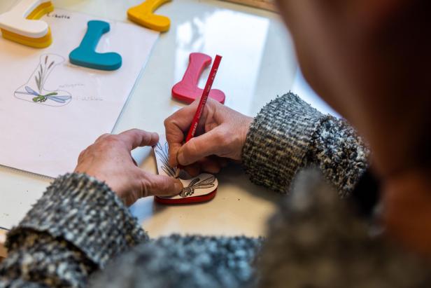 Eine Person zeichnet mit einem roten Stift eine Libelle auf einen roten Holzbuchstaben "L".
