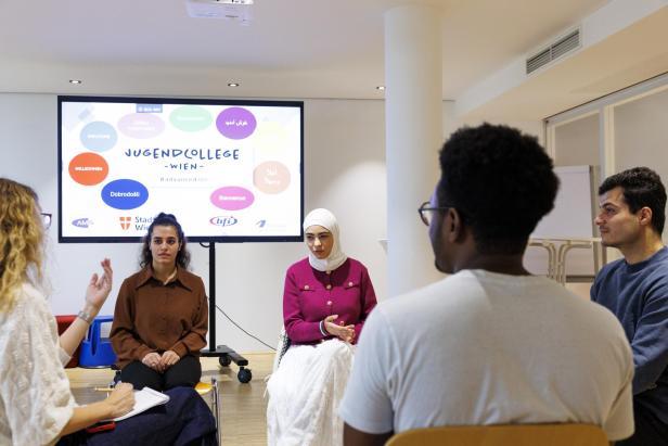 Eine Gruppe von Personen sitzt in einem Raum vor einer Leinwand des Jugendcollege Wien.