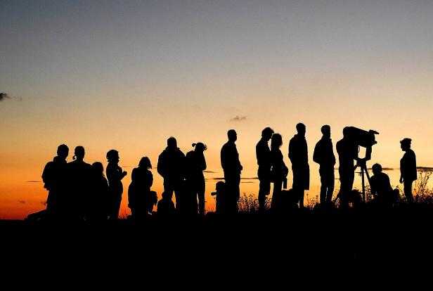 Silhouetten einer Gruppe von Menschen, einige stehen, einer blickt durch ein Teleskop, vor einem orangefarbenen Sonnenuntergangshimmel.