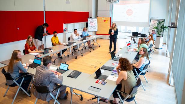 Auf dem Bild sieht man Menschen, die in einem Konferenzraum sitzen und einer Vortragenden zuhören.